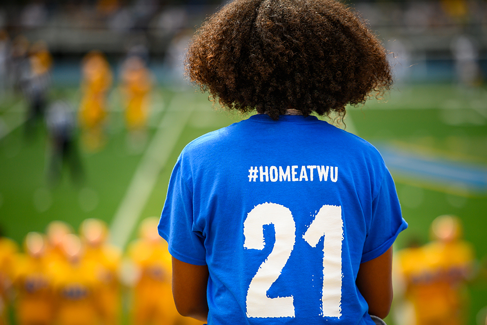 The back of a student wearing a blue shirt that says "#HomeAtWU 21" as they watch the football game
