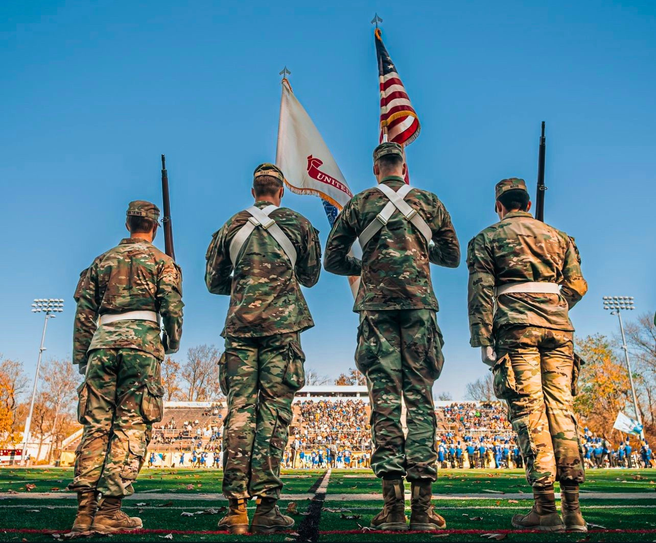 Four R-O-T-C cadets in fatigues, with their backs to the camera facing the stands in a stadium, hold flags and weapons as part of an honor guard