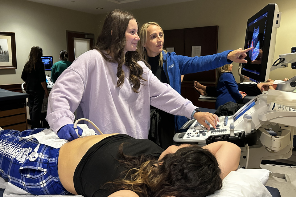 A student and instructor work at an ultrasound machine. Another student plays the role of patient.