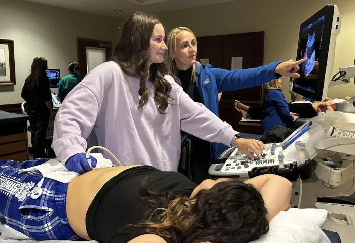 A student and instructor work at an ultrasound machine. Another student plays the role of patient.