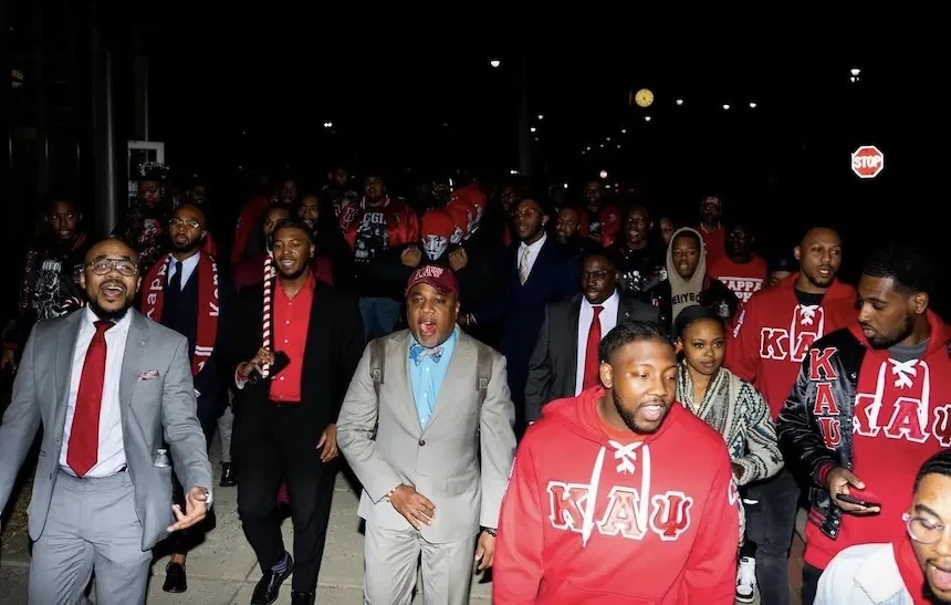 Large group of men wearing red Kappa Alpha Psi apparel walking together at night on a city sidewalk during a fraternity event.