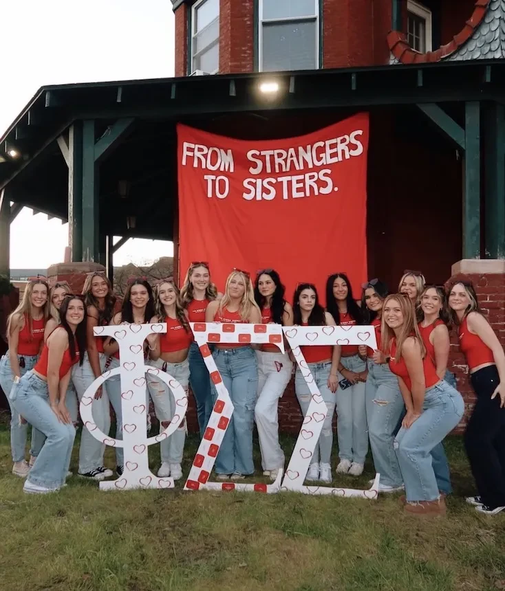 Group of Phi Sigma Sigma members posing outdoors in front of a large red banner reading “From Strangers to Sisters” while holding oversized Greek letters.