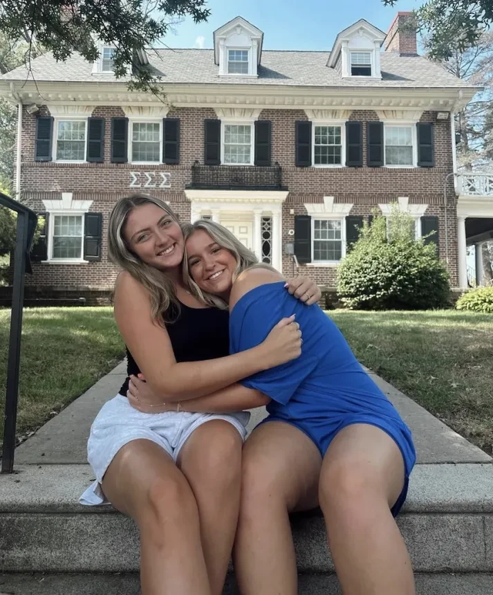 Two Sigma Sigma Sigma members sitting on steps hugging in front of a brick sorority house.