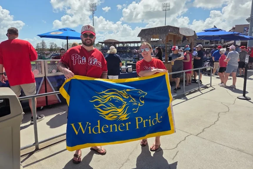 Alumni wearing Phillies gear pose for a photo with a blue and yellow "Widener Pride" flag
