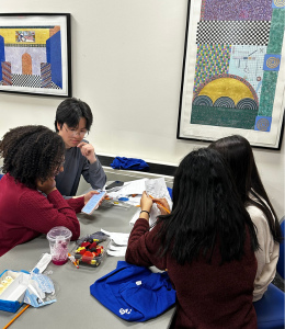Students sit at a table working to solve a mathematical puzzle. They are working with piece of paper and pencils, as well as their smartphones. 