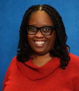 A headshot photo of Keeshea Turner Roberts. She smiles at the camera wearing a red top in front of a blue background.