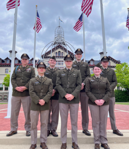 Six commissioining ROTC cadets and their lieutenant commander standing outdoors under American flags