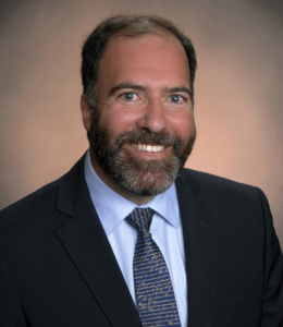 A headshot photo of Michael Dimino. He wears a dark suit, and is smiling at the camera. 