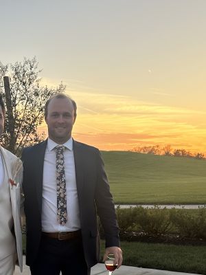 Cody Schulte poses for a photo wearing a suit. There is a colorful sunset behind him