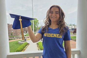 Amanda Rappa, wearing a blue Widener shirt and holding a blue grad cap, stands on the balcony of Old Main with campus behind her