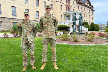 R-O-T-C cadets Liam Reilly and Braydn Erb, wearing Army fatigues, stand on the grass in front of Old Main with a statue in the background