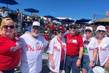 Alumni, friends and family pose for a photo at a Phillies spring training game. Everyone in this photo is wearing Phillies gear and smiling at the camera.