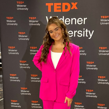 Amanda Rappa, in a pink pant suit, stands in front of a TedX backdrop