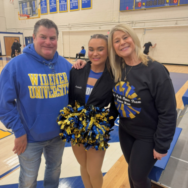 Amanda Rappa in her Pride Pack dance uniform holding blue and yellow pom poms, stands between her parents in the gym