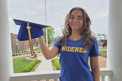 Amanda Rappa, wearing a blue Widener shirt and holding a blue grad cap, stands on the balcony of Old Main with campus behind her