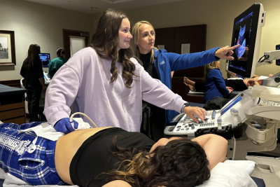 A student and instructor work at an ultrasound machine. Another student plays the role of patient.