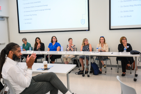 Seven professors sitting on a panel in front of a room, with an audience member clapping in the foreground