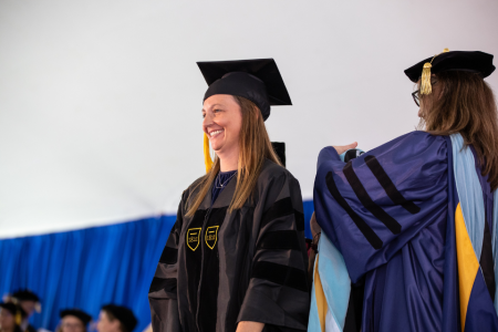 Tara Valoczki stands on stage in black Commencement robe and hat
