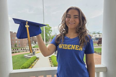 Amanda Rappa, wearing a blue Widener shirt and holding a blue grad cap, stands on the balcony of Old Main with campus behind her