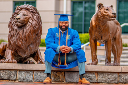 Delorean Andrews poses in his graduation cap and gown with the Pride of lions statue on campus.