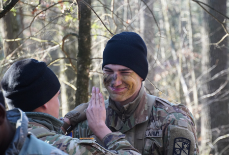 R-O-T-C cadet Braydn Erb in fatigues, face paint, and a hat high fives another cadet in the woods