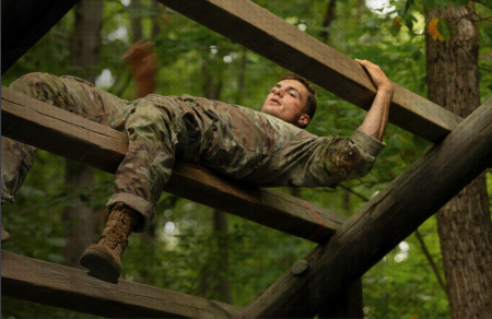 R-O-T-C cadet Liam Reilly, wearing Army fatigues, balances in the air on a wooden tree structure in the woods