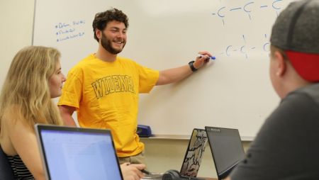 Three Widener University students study together for an upcoming exam, with one student standing at a whiteboard.
