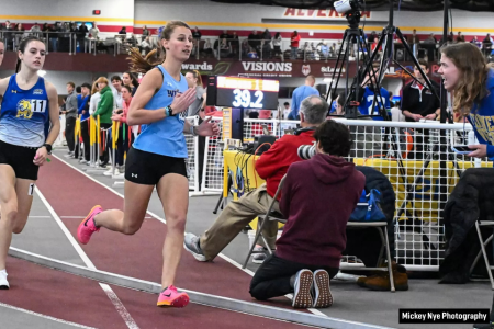 Student athlete Gabby Nye, in Widener track uniform, running on an indoor track with another runner behind her and people watching in the center of the oval