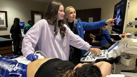 A student and instructor use an ultrasound machine. Another student plays the role of patient.
