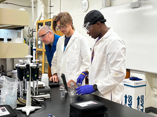 Two students in white lab coats and goggles work at a table with a professor in a lab