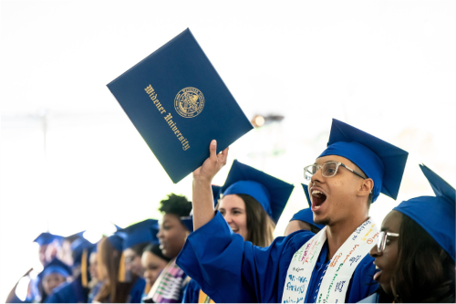 A student wearing a blue graduation cap and gown holds up his diploma case and cheers