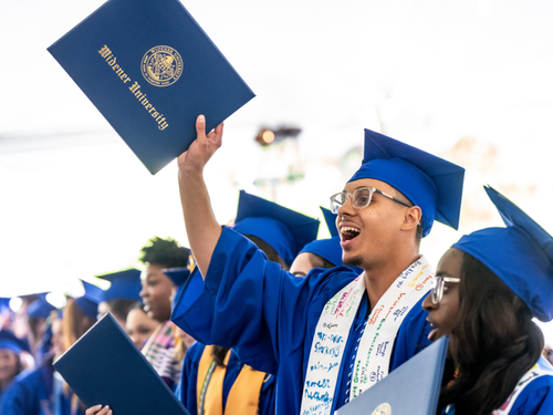 A graduate holds up his diploma, smiling and cheering.