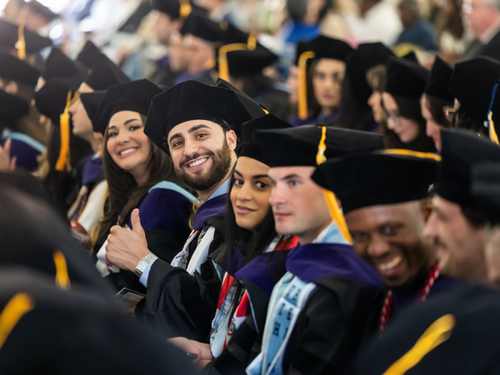 Graduates are seated in the crowd. One looks at the camera, smiling and giving a thumbs up.