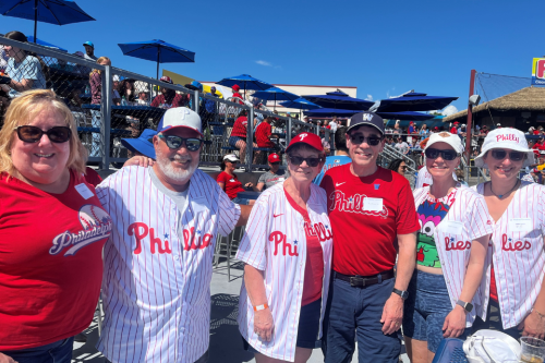Alumni, friends and family pose for a photo at a Phillies spring training game. Everyone in this photo is wearing Phillies gear and smiling at the camera.